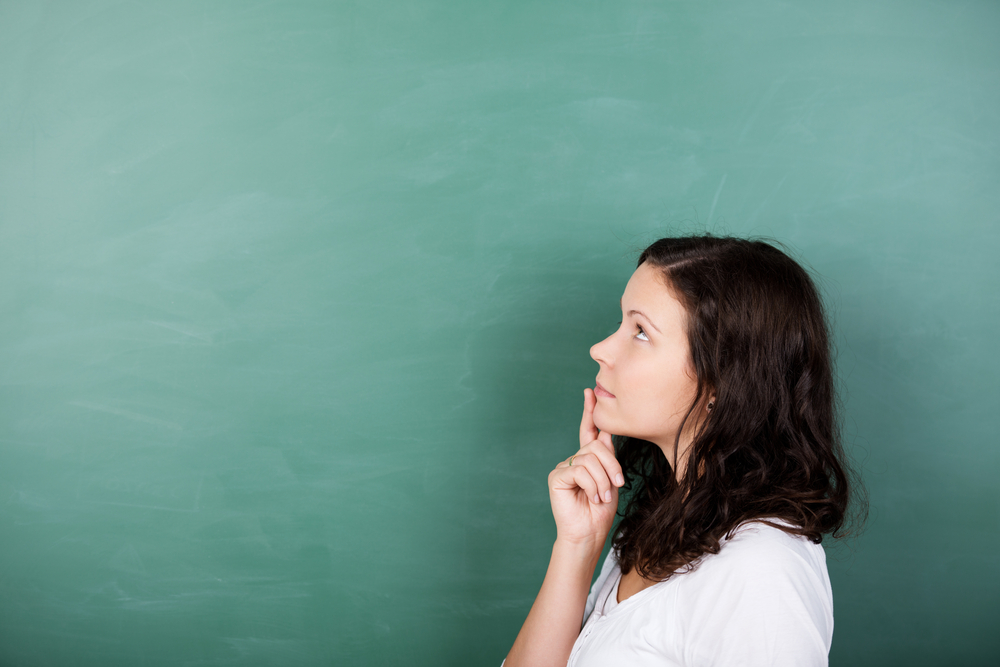 Attractive young female student standing solving a problem staring thoughtfully up into the air with her finger to her lips against a blank green blackboard with copyspace